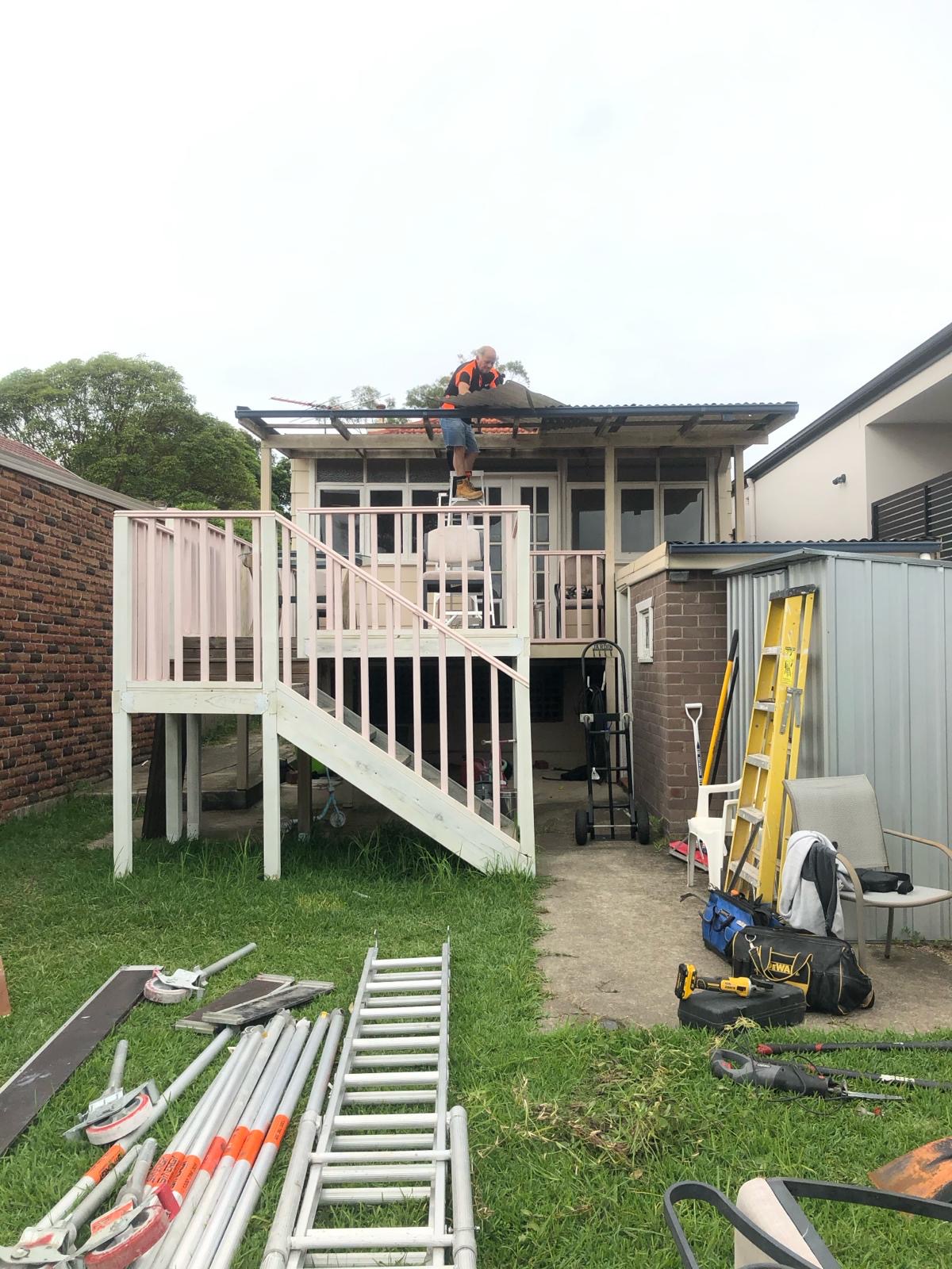 Raised rear deck and verandah before demolition — worker removing corrugated roof sheeting by hand